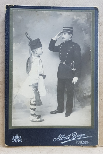 OFITER SALUTAND UN COPIL IMBRACAT IN COSTUM POPULAR CU PUSCA , BANDULIERA SI CUSMA , FOTOGRAFIE TIP CABINET , STUDIO ALBERT BOGA  - PLOIESTI , MONOCROMA, PE CARTON , CCA. 1900